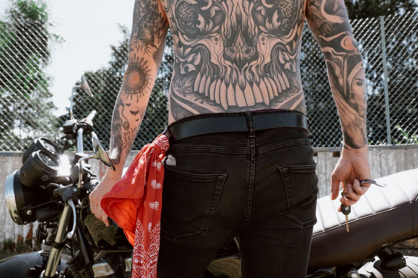 3d printed accessories for bike — Back view of a tattooed biker holding keys beside a motorcycle, showcasing detailed body art and black jeans