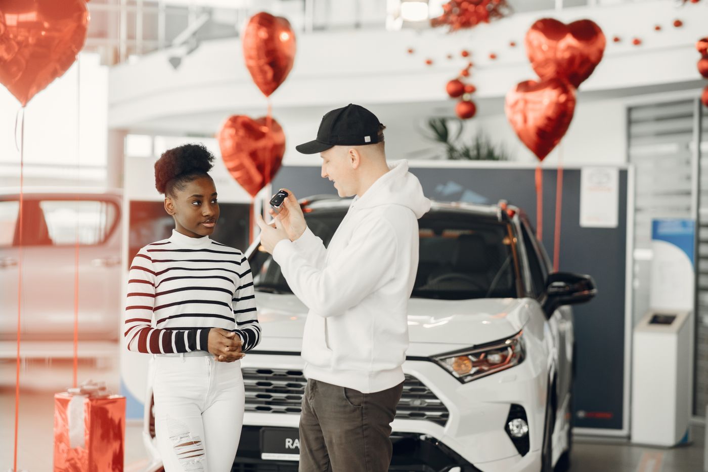personalised keychain for car — A couple exchanging car keys inside a showroom decorated with heart-shaped balloons, celebrating a new car purchase