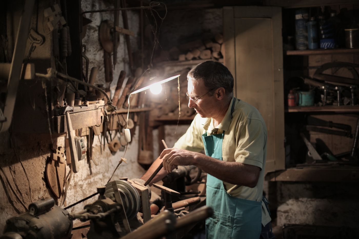 personalised keychains online india — Focused artisan in apron and eyeglasses using lathe machine while holding metal detail in hand during working in dark work