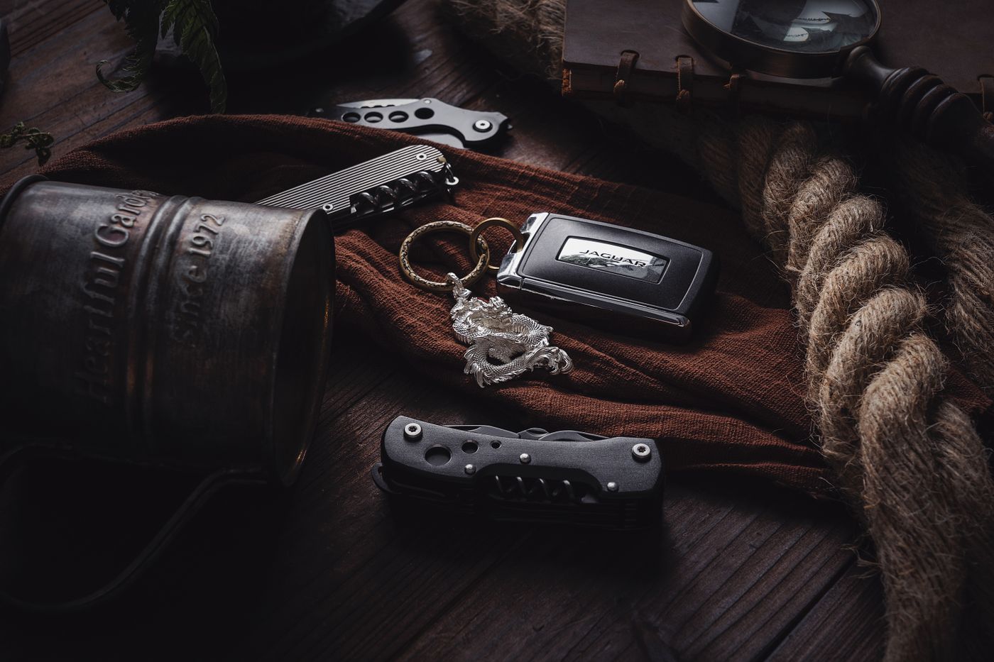anime keychain — A vintage-themed still life featuring a keychain, rope, knife, and an engraved mug on a wooden table