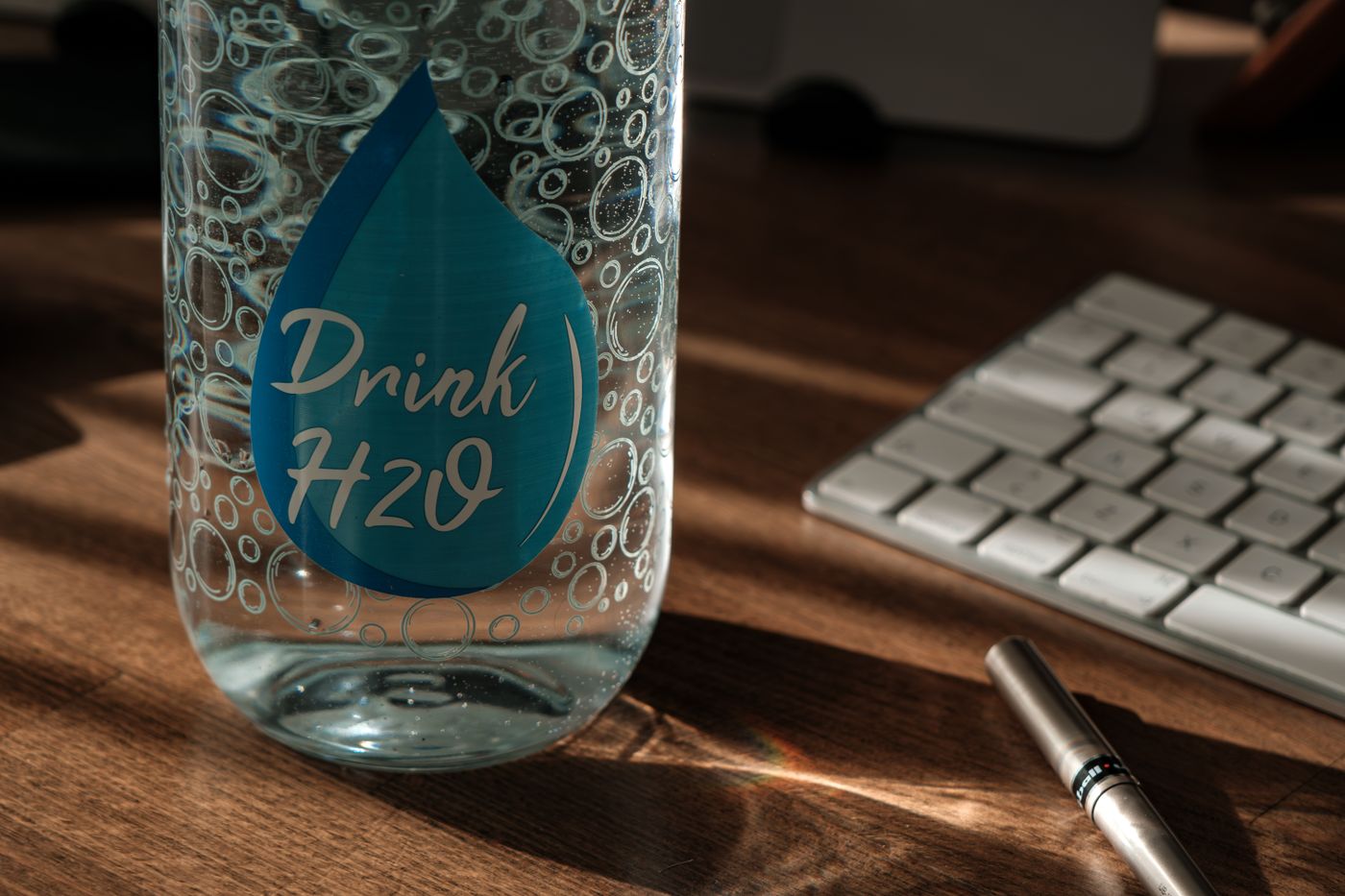 sticker for water bottle — Glass water bottle labeled 'Drink H2O' on an office desk with sunlight and keyboard